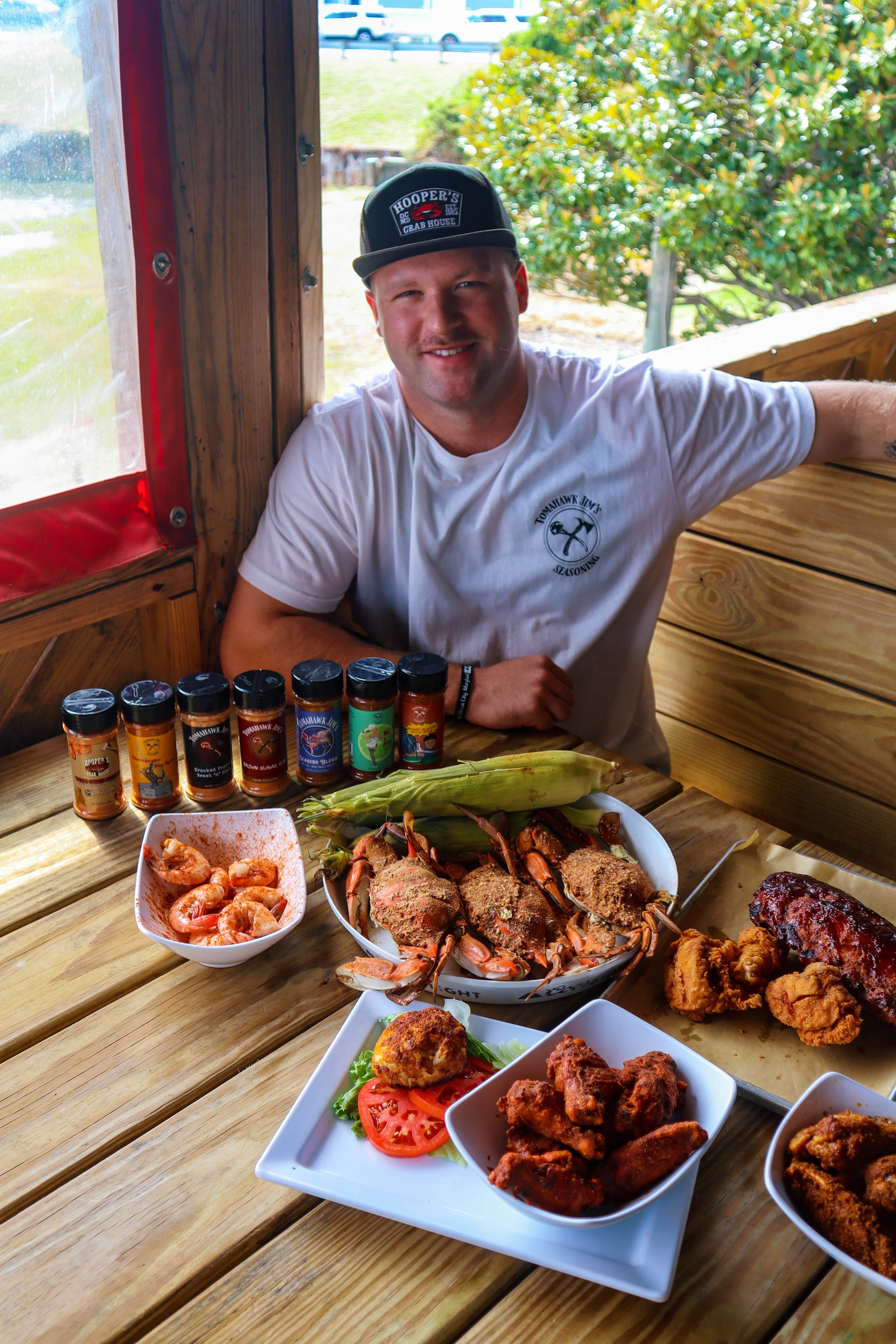 Man sitting at a wooden table with various food dishes and hot sauce bottles.